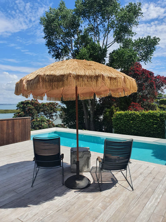 Hula 240cm raffia thatch market umbrella by pool with black chairs and blue sky background