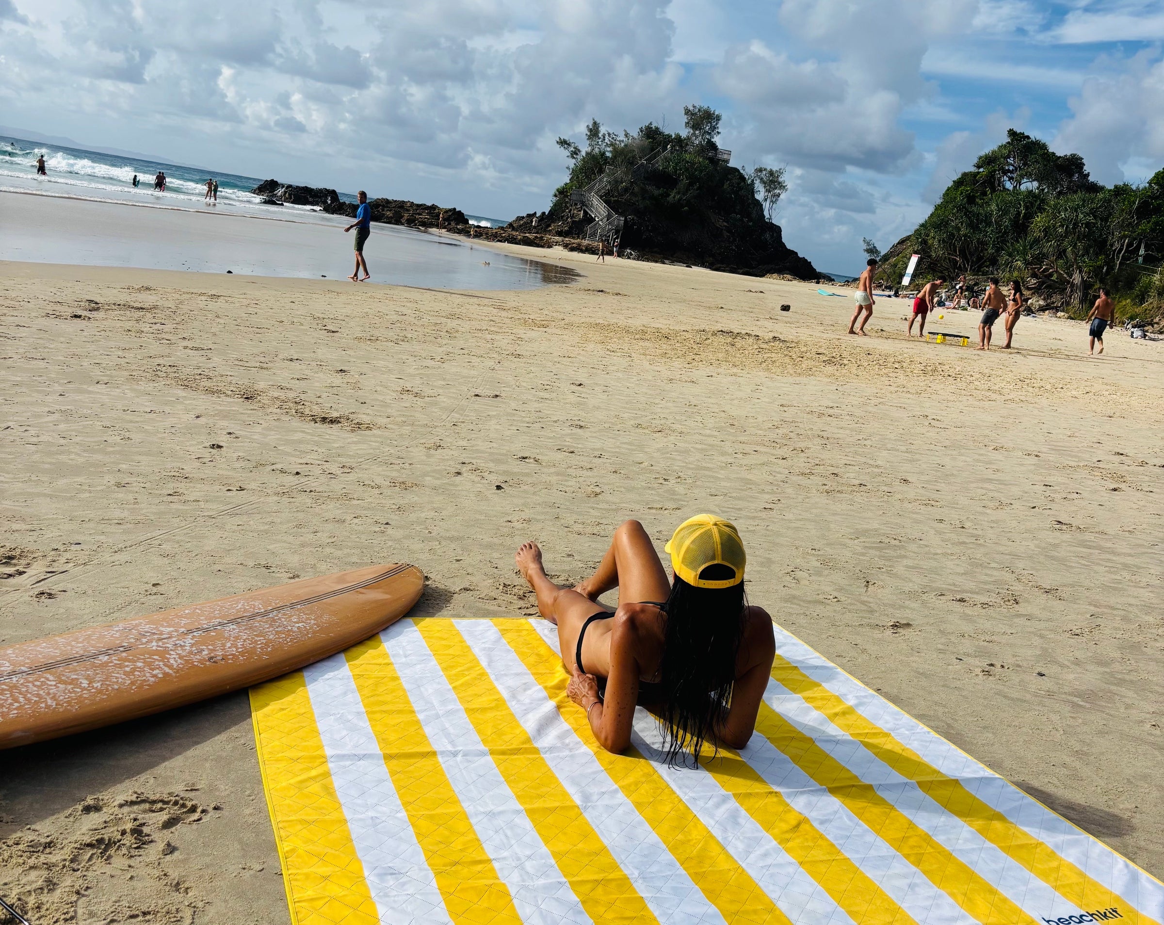 Person lying on a yellow and white striped towel at the beach with a surfboard