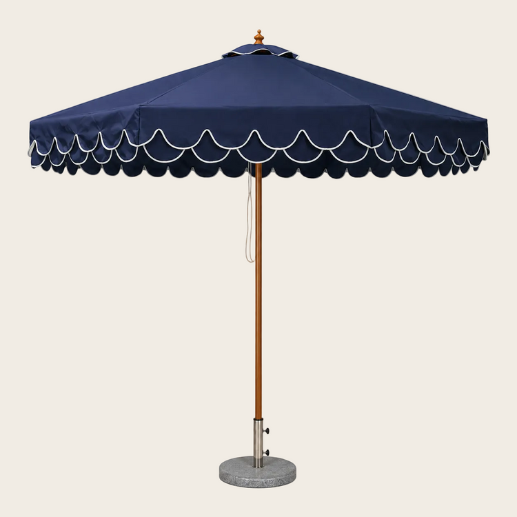 A navy blue patio umbrella with a scalloped white trim along the edge of the canopy, a brown wooden pole, and a round gray stone base on a white background.