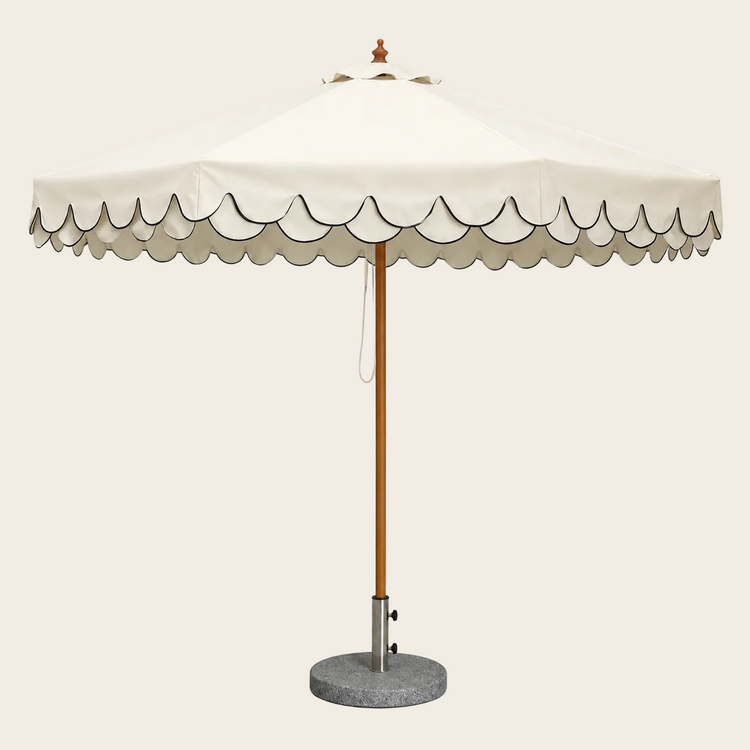 An outdoor patio umbrella with a cream-colored canopy featuring a scalloped edge, a natural wood pole, and a round gray concrete base on a white background.