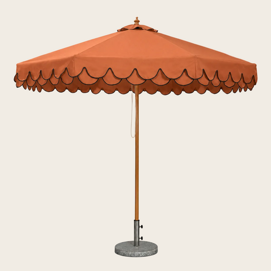 An orange patio umbrella with a scalloped edge, a wooden pole, and a round concrete base on a white background.